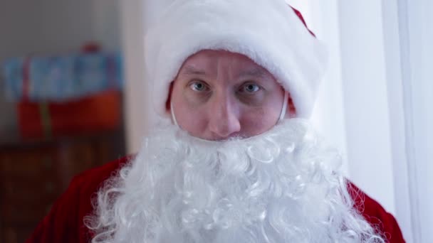 Kendine güvenen bir portre. Ciddi bir Noel Baba kameraya bakarak koronavirüs maskesi takıyor. Yakından bakınca, Noel Baba kostümü giymiş beyaz bir adam. Sakallı, Covid-19 yılbaşında. Kapalı alanda poz veriyor.. — Stok video