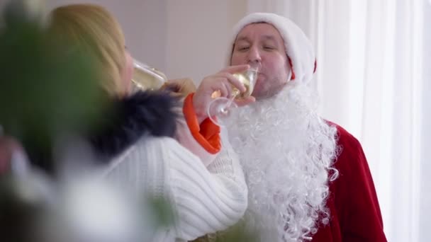 Retrato del hombre feliz en traje de Santa Claus bebiendo champán besándose mujer sonriendo. adulto caucásico marido en padre traje de Navidad celebrando vacaciones con esposa en casa. — Vídeos de Stock