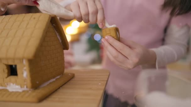 Live-Kamera verfolgt Ingwerkekse in Kinderhänden beim Auftragen von Zuckerleim, der Teig auf das Haus legt. Nicht wiederzuerkennendes kaukasisches Mädchen, das an Neujahr mit Mutter traditionelles süßes Dessert zubereitet. — Stockvideo