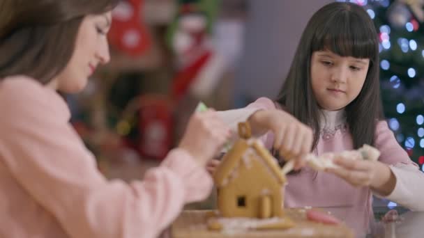 Koncentrerad söt vit flicka dekorera pepparkaksbakverk med kvinna på julafton inomhus. Porträtt av charmig dotter och mor gör traditionella ingefära hus på nyår hemma. — Stockvideo