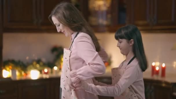 Dottern binder förkläde på leende mor som står i köket hemma. Medium sköt söt vit flicka hjälpa mamma matlagning på julafton inomhus. Familjeenhet och nyår. — Stockvideo