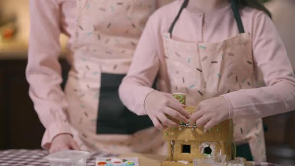 Live kamera går ner från leende liten flicka till pepparkakshus med händerna placera julgran söt dekoration. Glad barnmat Nyårsdessert med mamma hemma på semester. — Stockvideo