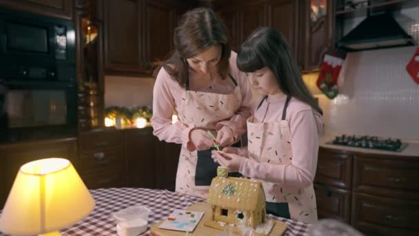 Приваблива кавказька дівчина поклала Санту на прикрашений імбирний будиночок, допомагаючи жінці готувати десерт увечері на кухні. Мила дочка і прекрасна мати роблять тістечка на Новий рік.. — стокове відео