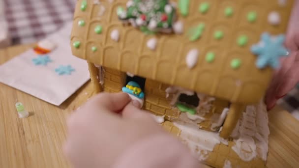 Gros plan fille méconnaissable plaçant bonbons bonhomme de neige sur la maison de pain d'épice. Petit enfant caucasien cuisine délicieux dessert sucré savoureux le soir de Noël à la maison. Nouvel An traditions concept. — Video