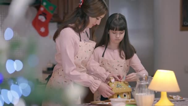Vit dotter hjälper mamma laga läckra pepparkakshus med julbelysning på framsidan. Porträtt av charmig flicka och vacker kvinna gör traditionella söta bakverk på nyår. — Stockvideo