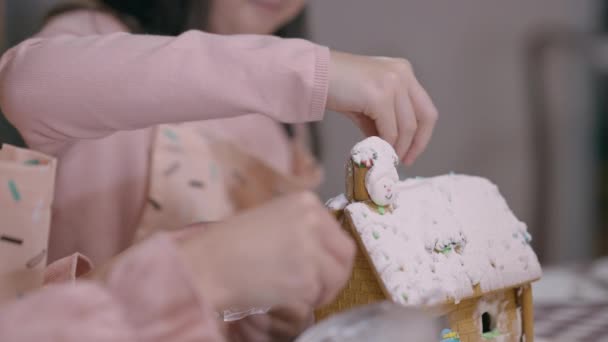 Filha branca ajudando a mãe a decorar a casa de gengibre na cozinha em casa na véspera de Ano Novo. Menina concentrada e mulher irreconhecível preparando sobremesa doce no Natal. — Vídeo de Stock