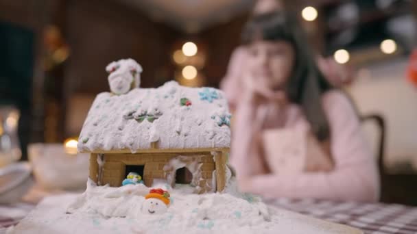 Pulveriserat dekorerat ingefära hus på bord med suddig kaukasisk mor och dotter kramas i bakgrunden. Glad kvinna och flicka matlagning på julafton njuter fritid hemma i köket. — Stockvideo