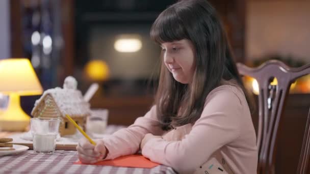 Pensativo muito caucasiano menina escrevendo carta para Papai Noel sentado à mesa em casa dentro de casa. Visão lateral retrato criança feliz desejando milagre na véspera de Natal. Ano Novo e conceito de infância. — Vídeo de Stock