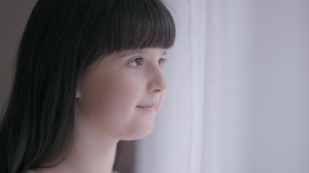 Close-up side view cute Caucasian girl looking out the window smiling at home indoors. Happy pretty brunette child with brown eyes enjoying weekend leisure. Childhood concept. — Stock Video