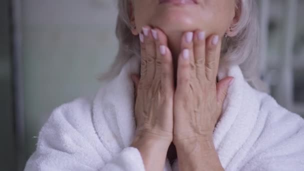 Primer plano del cuello de la mujer madura masajeando la piel con las manos por la mañana. Vista frontal confiada señora de edad caucásica cuidando de la belleza natural en el interior en el baño en casa. — Vídeo de stock