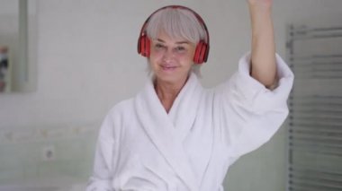 Ön manzara portresi, kulaklıklı neşeli olgun bir kadın banyoda yavaş çekimde dans ediyor kameraya bakıp gülümsüyor. Kafkasyalı orta yaşlı bayanın sabah hobisinin keyfini çıkarın..