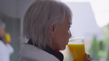 Yakın çekim pozitif kaygısız orta yaşlı beyaz kadın evde pencereden dışarı bakarak portakal suyunun tadını çıkarıyor. Kendine güvenen olgun bir kadının hafta sonu sabahı kutlu olsun..