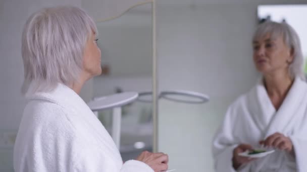 Confident beautiful middle aged Caucasian woman applying cucumber on eye skin reflecting in mirror in bathroom. Mature grey-haired attractive lady taking care of face in the morning. — Stock Video