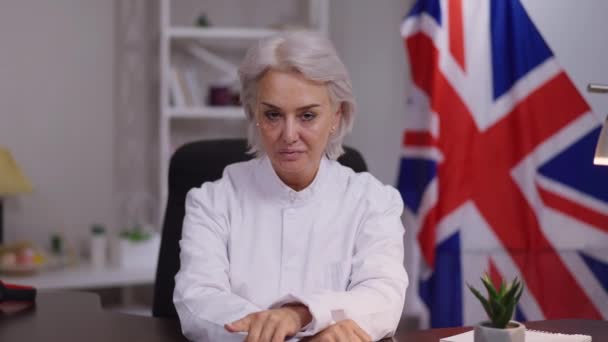 Retrato de una seria mujer de negocios madura profesional escuchando cuidadosamente sentada en el interior de la oficina con la bandera británica en segundo plano. Video chat POV de gestora exitosa conferencia en línea. — Vídeo de stock