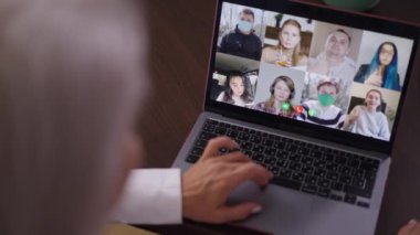 Yüksek açılı görüntülü laptop, görüntülü sohbet ve gri saçlı iş kadını parmaklarını yavaş çekimde hareket ettiriyor. Tanımlanamayan orta yaşlı beyaz yönetici çalışanlarla online beyin fırtınası yapıyor..