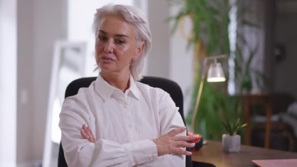 Femme mature fière confiante croisant les mains au ralenti en regardant la caméra assise dans le bureau à l'intérieur. Portrait d'attrayant mince PDG caucasien posant sur le lieu de travail avec une expression faciale sérieuse. — Video