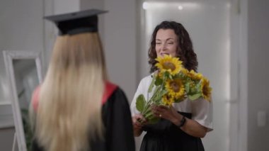 Gülümseyen mutlu anne, kızının konuşmasına bir buket sarı ayçiçeği veriyor. Mezuniyet gününde lise öğrencisini çiçeklerle kutlayan neşeli beyaz kadın portresi..