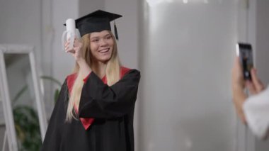 Heyecanlı mezuniyet kızı, sarılmış diplomayla evde akıllı telefondan fotoğraf çeken bir anne gibi poz veriyor. Mutlu, kendinden emin, gülümseyen, diplomalı bir kadının portresi..