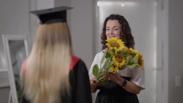 Smiling happy proud mother giving bouquet of yellow sunflower to graduate daughter talking. Portrait of cheerful Caucasian woman congratulating high school student on graduation day with flowers. — Stock Video