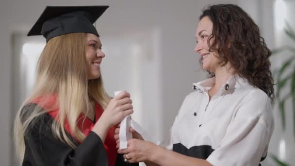 Sonriendo feliz madre e hija sosteniendo certificado rodado mirando el uno al otro de pie en el interior. Alegre mujer caucásica y graduado disfrutando del día de graduación en casa con diploma. — Vídeos de Stock