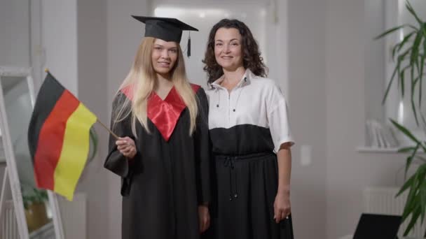 Mittleres Portrait glückliche kaukasische Mutter und Tochter im Abschlussoutfit posieren mit deutscher Fahne zu Hause. Glückliche selbstbewusste Frau und Absolventin, die in die Kamera lächelt. Stolz und Bildung. — Stockvideo
