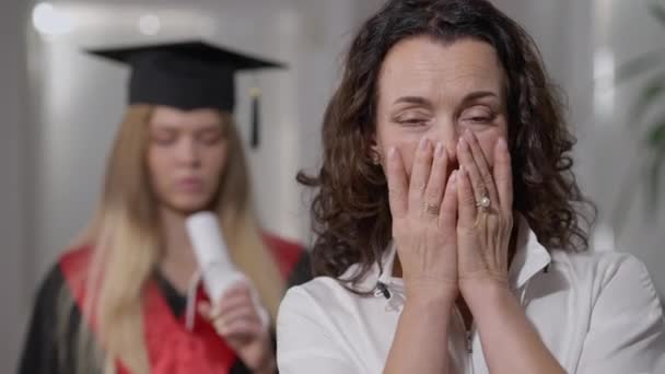 Maturo donna caucasica piangendo come figlia laureata offuscata in abito e cappello in piedi sullo sfondo. Ritratto di madre ansiosa preoccupata con giovane laureata a casa al chiuso. Ansia e concetto di graduazione. — Video Stock