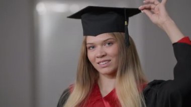 Mezuniyet elbisesi ve solda şapka tasmasıyla poz veren mutlu, kendine güvenen bir kadın portresi. Gururlu, zeki, beyaz, genç bir bayan mezun kameraya bakıp gülümsüyor..