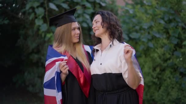 幸せな成熟した母親と若い娘が卒業日に屋外でポーズをとって英国の国旗に包まれました。公園に立って笑顔カメラを見て自信を持って白人女性の肖像画。スローモーション. — ストック動画