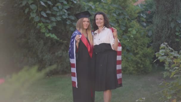 Широкий снимал веселую мать и дочь в выпускном платье, позируя с американским флагом, размахивающим в замедленной съемке. Счастливые кавказки, стоящие на летнем весеннем заднем дворе и улыбающиеся в камеру. — стоковое видео