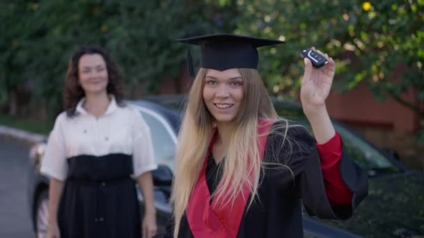 Proud graduate woman boasting car key standing outdoors with blurred happy mother at background. Portrait of wealthy Caucasian young beautiful grad student posing at new vehicle showing off. — Stock Video