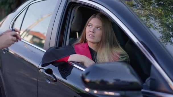 Charmig smal kvinna i examensrock sitter i bilen som oigenkännlig förälder passerar nyckel i slow motion. Porträtt av överraskad glad vacker kaukasisk grad ta emot fordon som gåva leende. — Stockvideo