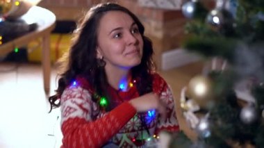 Neşeli, genç, zayıf bir kadın Noel ağacını evde Noel ışıklarıyla süslüyor. Heyecanlı mutlu beyaz kadın portresi. Oturma odasında tatilin tadını çıkarıyor..