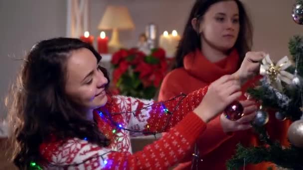 Mooie jonge gelukkige vrouw versieren kerstboom met vriend binnen. Portret van vrolijke blanke vrouwen die genieten van nieuwjaarsvoorbereidingen in de woonkamer. Feestdagen concept. — Stockvideo