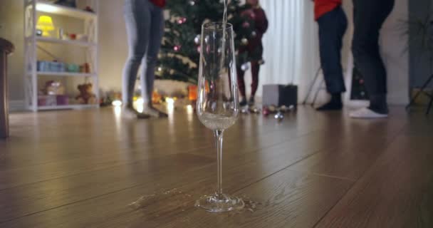 Szklanka z bliska z szampanem wylewającym się na podłogę i zamazanymi ludźmi dekorującymi choinkę na tle. Koncepcja noworocznych tradycji i przygotowań. Kino 4k ProRes kwatera główna. — Wideo stockowe