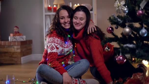 Dos jóvenes mujeres caucásicas felices sentadas en el árbol de Navidad abrazándose sonriendo mirando para otro lado hablando. Positivo hermosas amigas milenarias celebrando Año Nuevo en el interior de la sala de estar. — Vídeos de Stock
