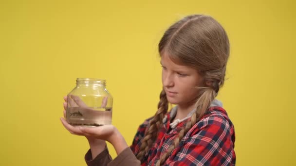 Widok z boku szczupła nastolatka trzyma butelkę z rybami stojącymi na żółtym tle. Portret pięknej, pewnej siebie, białej nastolatki podziwiającej małego suma. Koncepcja zwierząt i ludzi. — Wideo stockowe