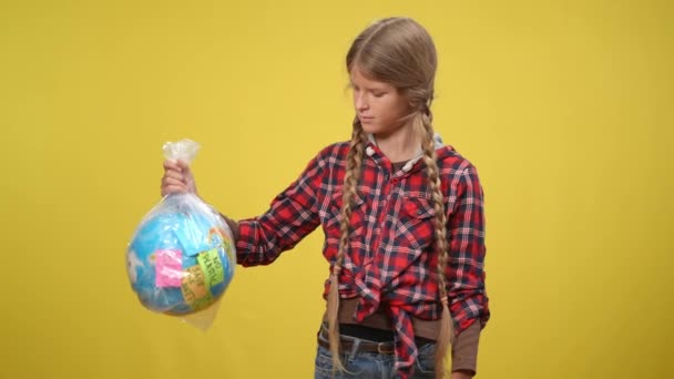 Střední záběr vážné dospívající dívka pózovat na žlutém pozadí s zeměkoulí s eko zprávy potřásající hlavou ne. Žena kavkazský teenager drží symbol planety v plastovém sáčku při pohledu do kamery. — Stock video