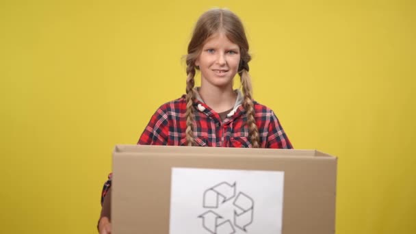 Улыбающаяся девочка-подросток держит картонную коробку с символом вторичной переработки, смотрящей в камеру. Портрет дружелюбного уверенного кавказского подростка позирующего на желтом фоне. Концепция экологических проблем. — стоковое видео