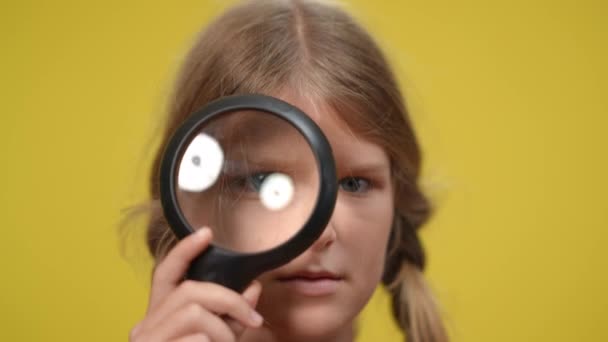 Faccia da primo piano di ragazza adolescente con occhio in lente di ingrandimento a sfondo giallo. Concentrato grave caucasico adolescente posa guardando la fotocamera. — Video Stock