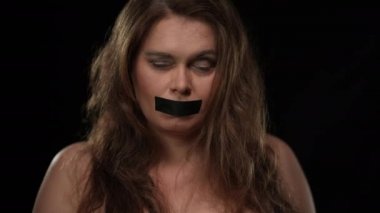 Yakın plan ve beyaz kadın kameraya bakarak ağızdan bandı yırtıyor. Siyah arka planda poz veren kendine güvenen esmer kadın. Dilsizlik ve özgürlük kavramı.
