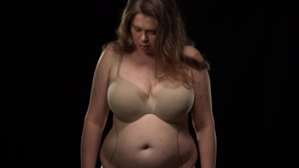 Pedestal shot overweight obese Caucasian woman in bra with marks for plastic operation at black background. Portrait of plus size sad lady looking at camera posing in underwear. Beauty standard. — Stockvideo