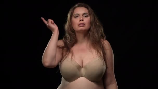 Self assured plus size Caucasian woman looking at camera gesturing think gesture. Portrait of confident overweight lady in bra touching temple with finger posing at black background. — 图库视频影像