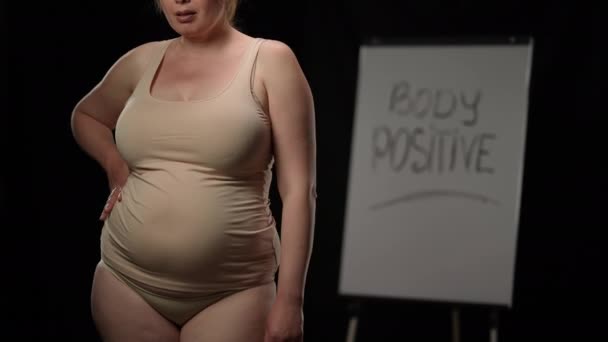 Unrecognizable plus size woman with hand on hip gesturing thumb up posing at black background with Body positive on whiteboard. Diversity and tolerance concept. — Vídeo de Stock