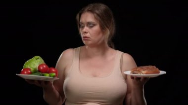 Sağlıksız tatlı seçen şişman kadının portresi siyah arka planda sağlıklı sebzelere karşı. Güzel, beyaz tenli, iri yarı, nefis kokan, gülümseyen bir kadın. Diyet molası.