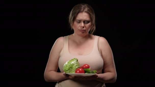 รูปภาพเศร้า Caucasian บวกขนาดผู้หญิงที่มีแผ่นผักในพื้นหลังสีดํา อารมณ์เสียผู้หญิงที่มีน้ําหนักเกินโพสกับอาหารมังสวิรัติเพื่อสุขภาพที่มองกล้อง โรคอ้วนและอาหาร . — วีดีโอสต็อก