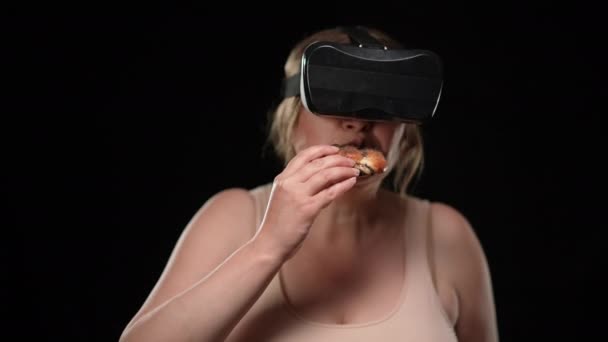 Избыточный вес белая женщина в гарнитуре VR едят вкусные нездоровые булочки на черном фоне. Портрет ожирённой леди, наслаждающейся нарушающей реальность диетой. Современные технологии и ожирение. — стоковое видео