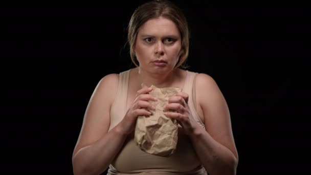 Front view scared Caucasian plus size woman with frightened facial expression breathing in paper bag posing at black background. Overweight lady posing having panic attack. — Stockvideo
