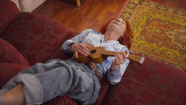 High angle view carefree retro woman singing playing ukulele lying on cozy couch indoors. Happy cheerful Caucasian redhead lady enjoying weekend leisure and hobby at home. Slow motion. — Vídeo de Stock