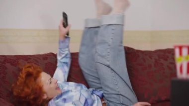 Joyful retro woman jumping on sofa in slow motion laughing turning on TV with remote control. Portrait of relaxed cheerful Caucasian lady having fun resting at home on weekend. Lifestyle and leisure.
