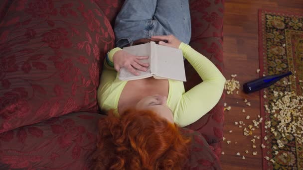 Top view κοκκινομάλλα ρετρό γυναίκα κοιμάται με το βιβλίο και άδειο μπουκάλι μπύρας που βρίσκεται στο πάτωμα με διάσπαρτα ποπ κορν. Μεθυσμένη εξαντλημένη Καυκάσια κυρία κοιμάται στο σπίτι σε άνετο καναπέ σε εσωτερικούς χώρους. — Αρχείο Βίντεο